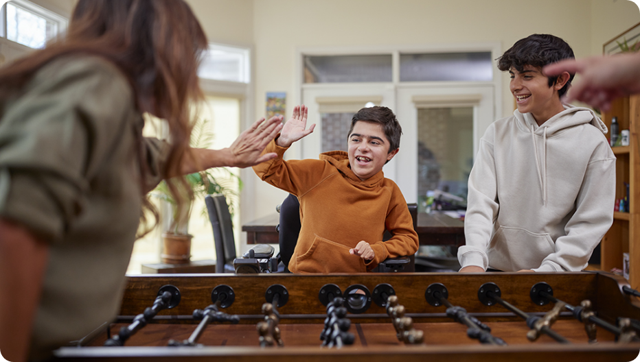 A VILTEPSO patient in an orange hoodie high-fiving a friend while playing foosball in a cheerful living space.