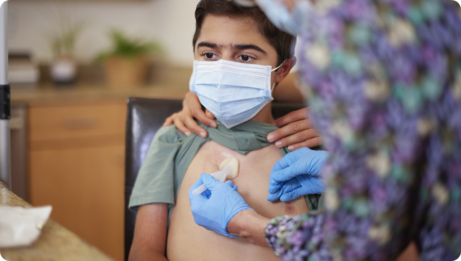 Masked VILTEPSO patient receiving port site care from a gloved caregiver in a clinical home setting.