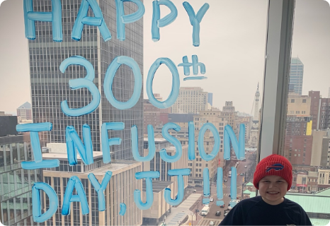 Jordan wearing a red hat, smiling in front of a window with 'Happy 300th Infusion Day, JJ!!!' written in blue letters on the glass.